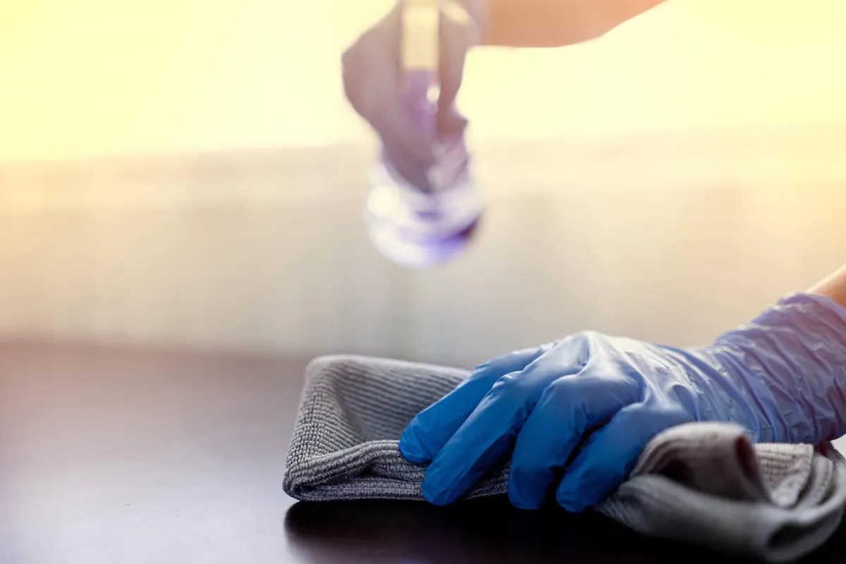 Eine Person mit blauen Handschuhen reinigt eine Oberfläche mit einem Tuch und sprüht Reinigungsmittel darauf.