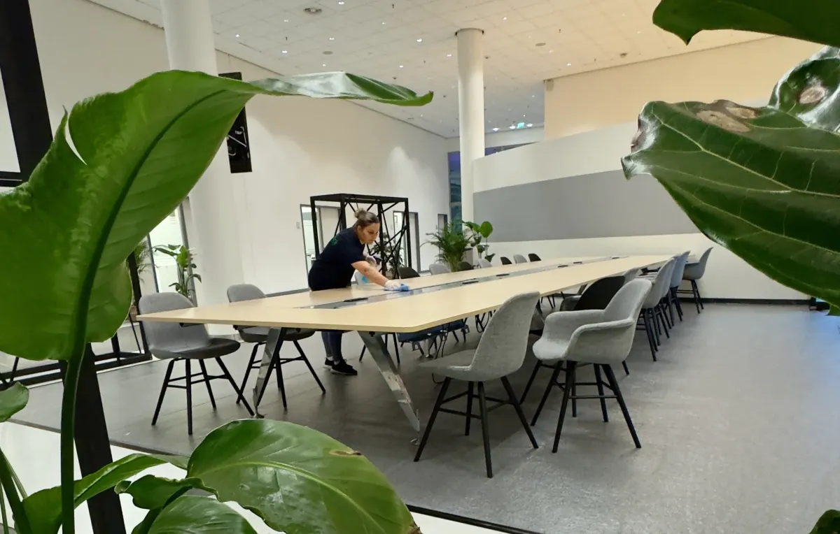 Eine Person reinigt einen langen Konferenztisch in einem modernen Besprechungsraum mit grauen Stühlen und grünen Pflanzen.