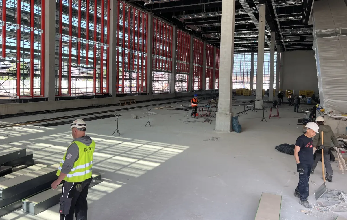 Bauarbeiter in Schutzkleidung arbeiten bei Tageslicht in einem Gebäude mit großen Fenstern und freiliegenden Deckenbalken.