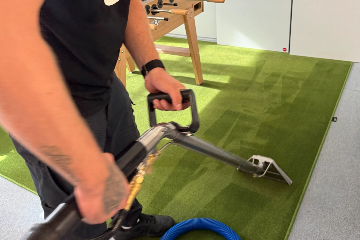Eine Person reinigt einen grünen Teppich mit einer Teppichreinigungsmaschine neben einem Tischfußballtisch.