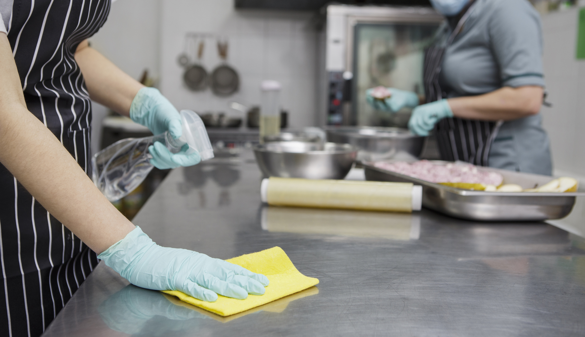Zwei Personen mit Handschuhen und Schürzen reinigen eine Großküche und bereiten auf einer Edelstahltheke Essen zu.