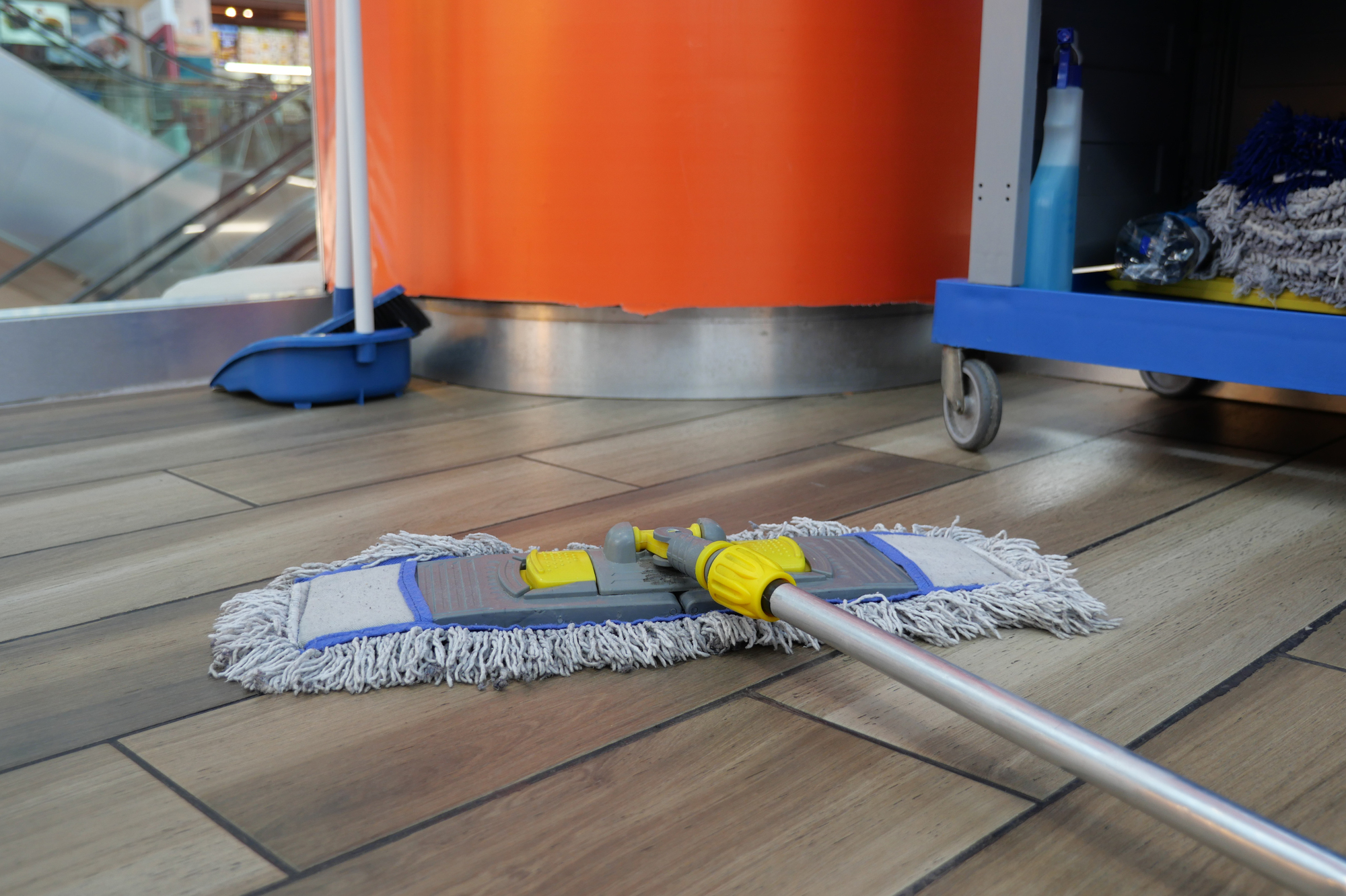 Ein Flachmopp mit gelbem Griff liegt zur Bodenreinigung auf einem Holzboden neben Reinigungsmitteln vor einer orangefarbenen Wand.