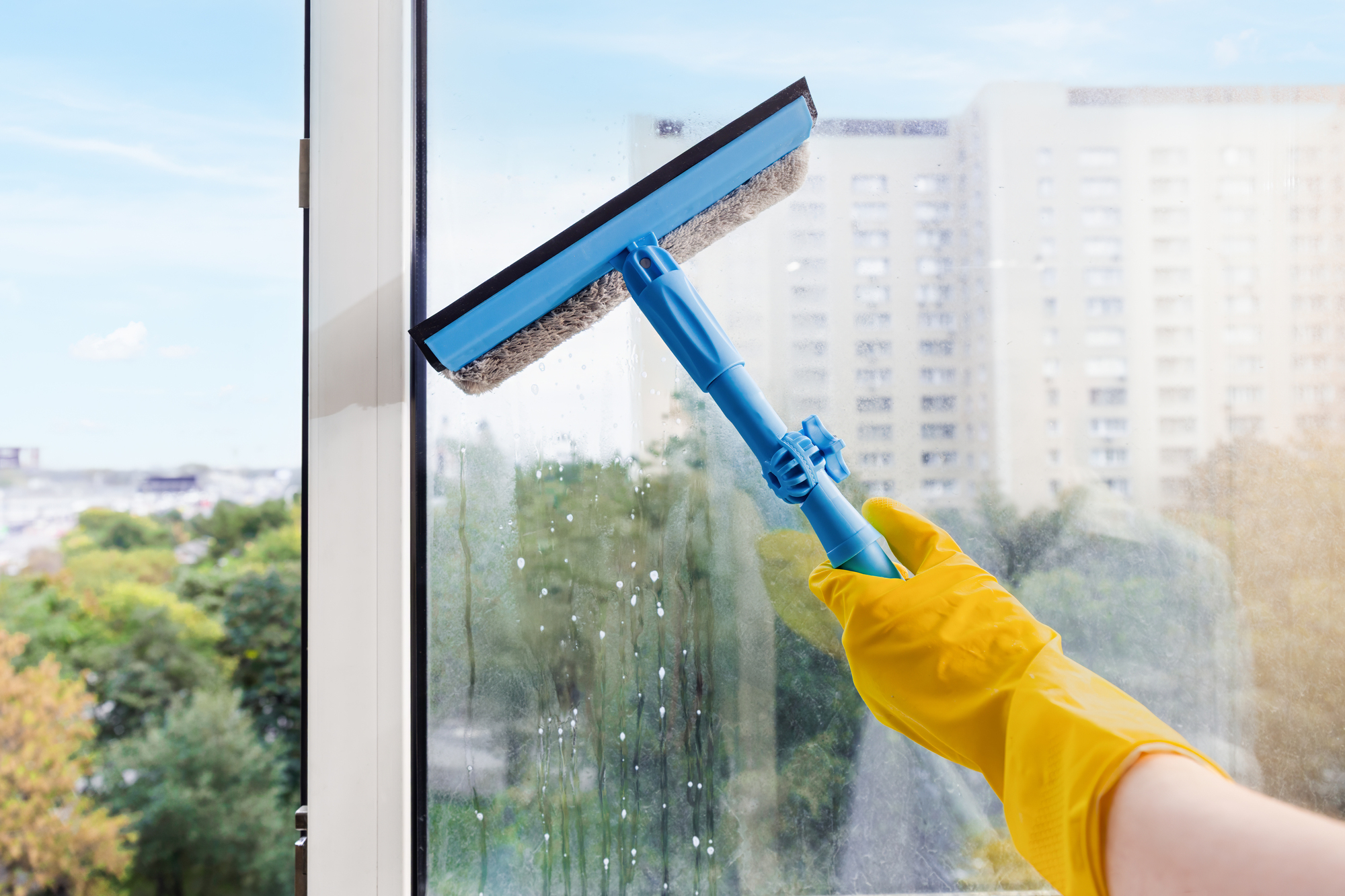 Eine Person reinigt Fenster mit einem blauen Abzieher und einem gelben Handschuh; im Hintergrund sind Gebäude und Bäume zu erkennen.