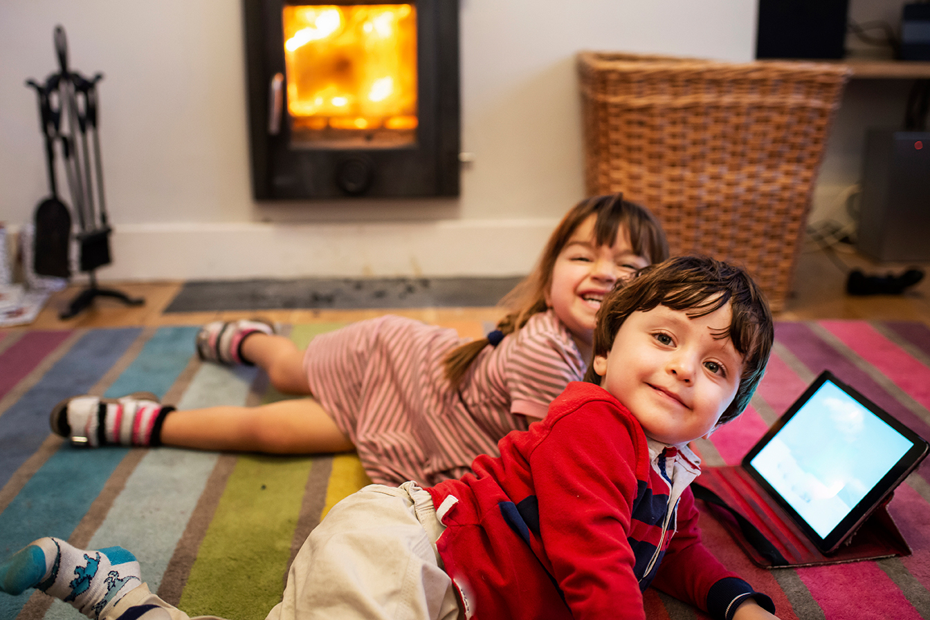 Zwei Kinder liegen lächelnd auf einem gestreiften Teppich vor dem Kamin und schauen gemeinsam auf ein Tablet.