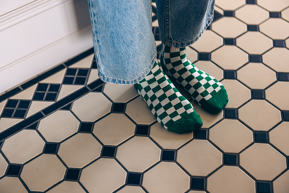 Eine Person mit grün-weiß karierten Socken und blauen Jeans steht auf einem schwarz-weiß gefliesten Boden.