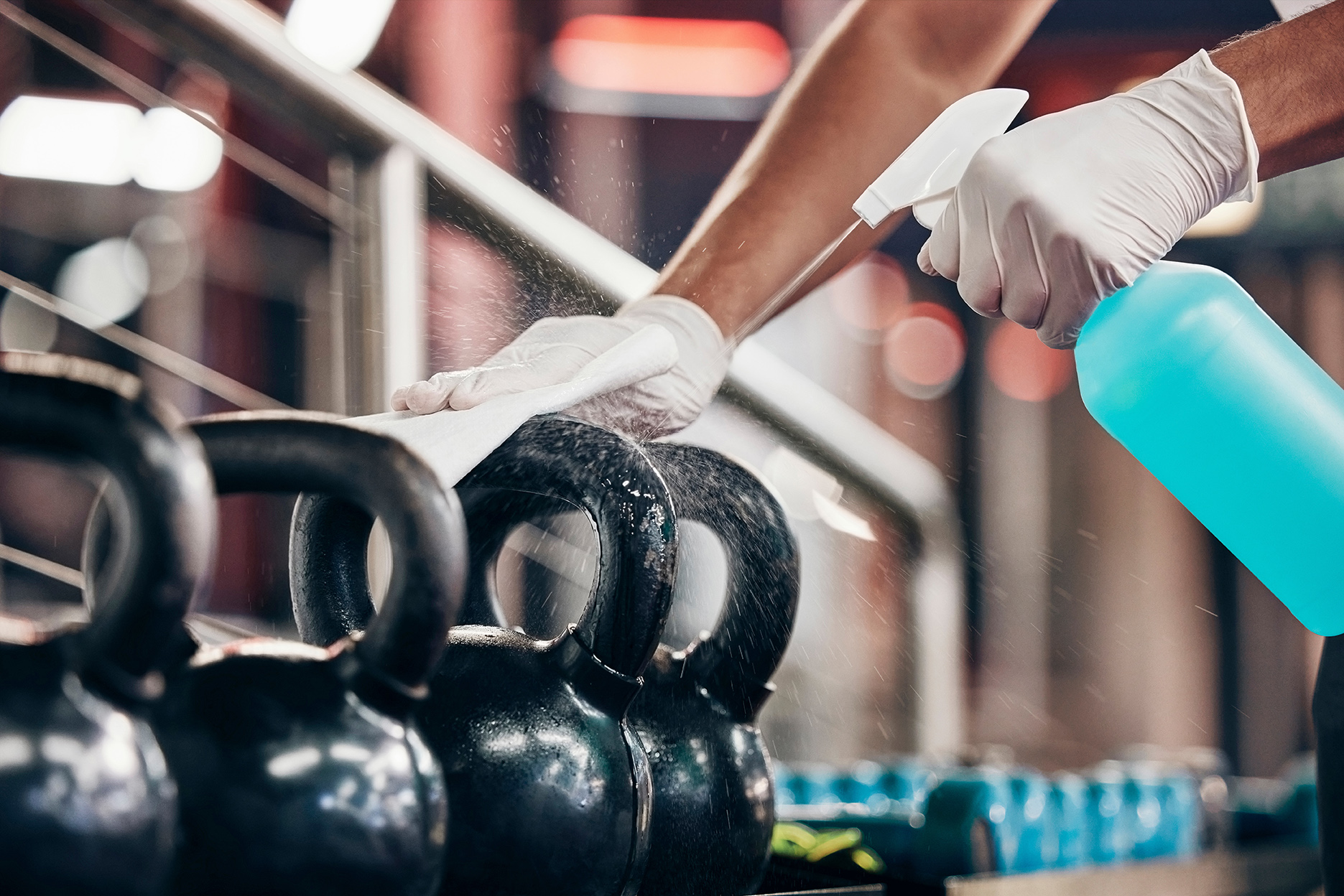 Eine Person reinigt im Fitnessstudio schwarze Kettlebells mit einem Tuch und einer blauen Sprühflasche und trägt dabei Handschuhe.