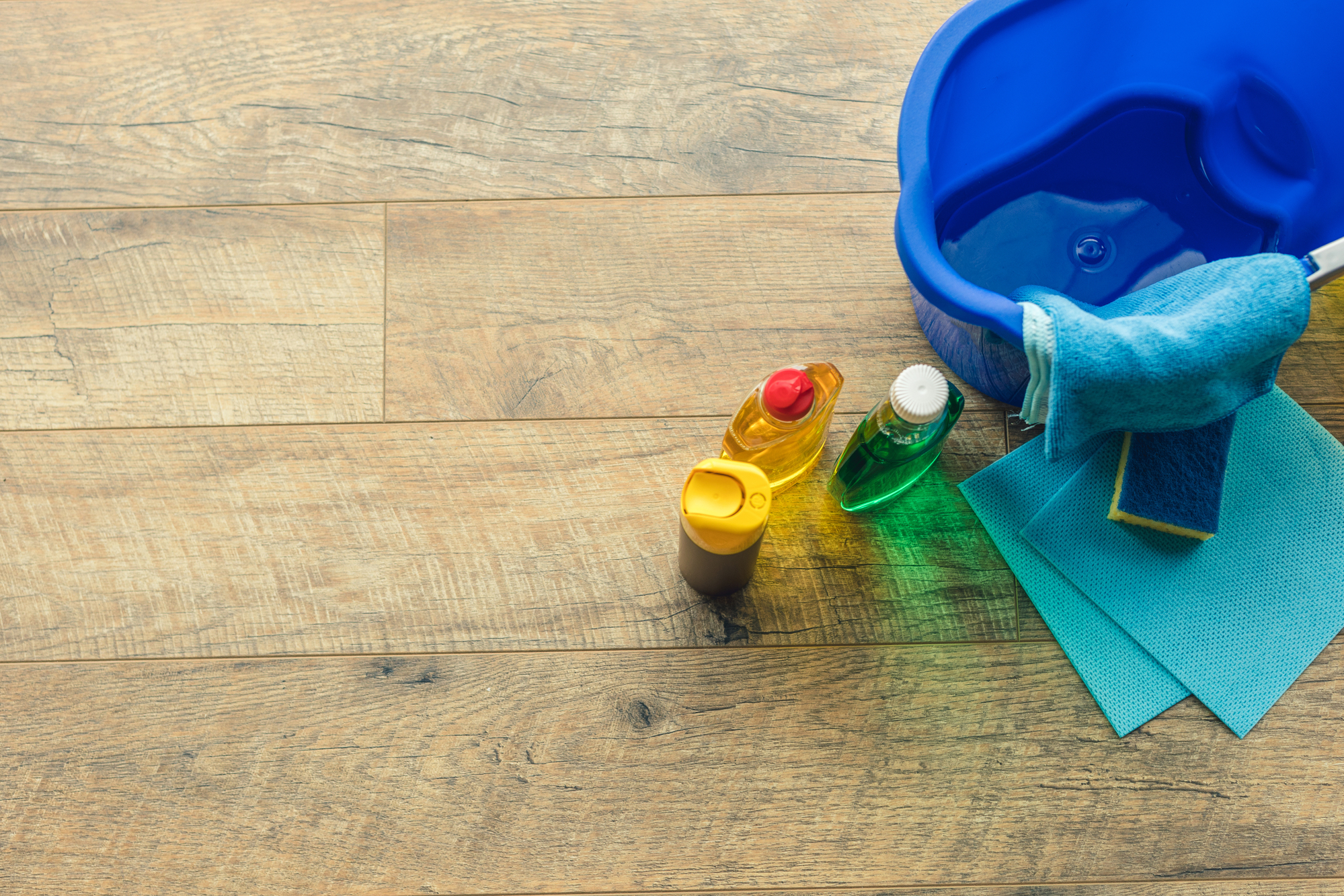 Reinigungsutensilien wie ein blauer Eimer mit Wasser, ein Wischmopp, Reinigungsmittel, ein Schwamm und Tücher auf einem Holzboden.