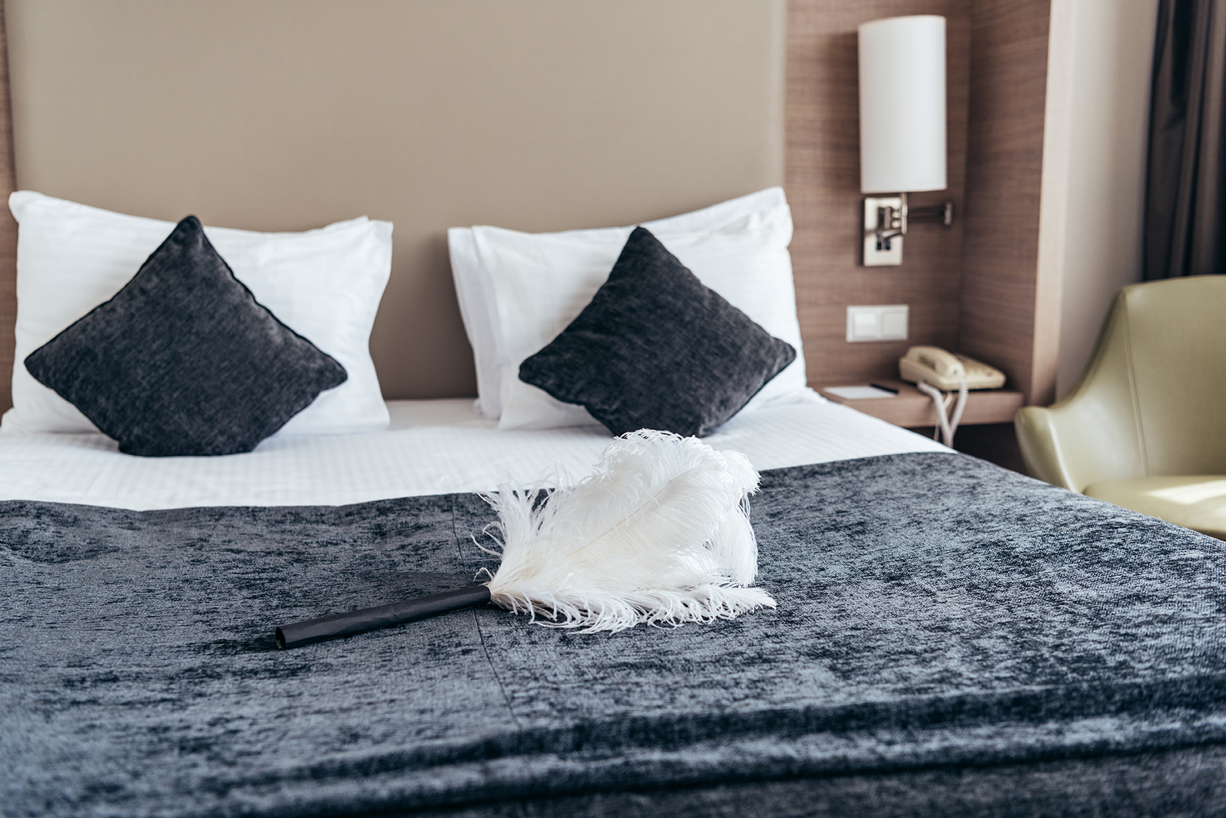Ein Bett mit weißen Kissen, dunklen Polstern, einer Decke, einem Staubwedel, einem Telefon und einem grünen Stuhl in einem Zimmer.